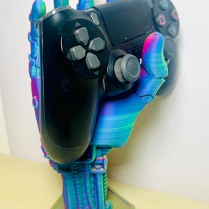 May include: A 3D printed hand-shaped stand with a colorful, iridescent finish holds a black PlayStation 4 controller. The stand has a hexagonal base.