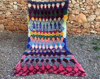 Può includere: Un vivace tappeto tessuto a mano con un motivo geometrico a rombi. Il tappeto presenta una ricca tavolozza di colori, tra cui rosa, blu, verde, giallo e viola. Il tappeto è realizzato in materiale spesso e strutturato ed è adatto per un soggiorno o una camera da letto.