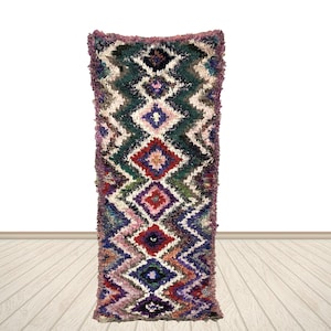 May include: A rectangular, multi-colored rug with a geometric diamond pattern. The rug features a mix of colors including green, red, purple, and white. The rug is displayed against a white background and a wooden floor.