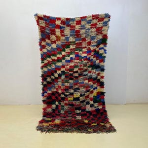 May include: A rectangular, hand-woven rug with a colorful checkered pattern. The rug features a mix of red, blue, green, yellow, and cream squares. The rug has a shaggy texture and is displayed against a white wall.