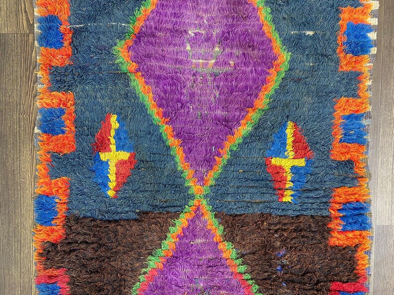 May include: A vibrantly colored, hand-woven rug with a geometric design. The rug features a central purple diamond shape, framed by orange and green, set against a blue background. Accents include red, yellow, and blue diamond shapes. The rug's edges are trimmed with orange.