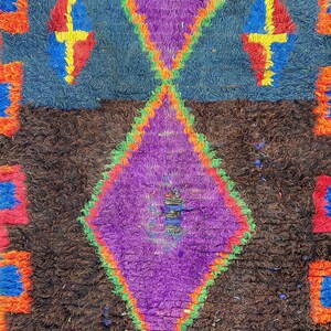 May include: A colorful, textured rug with a geometric design. The rug features a central purple diamond shape, framed by green and orange, set against a brown background. Blue and orange accents complete the design.