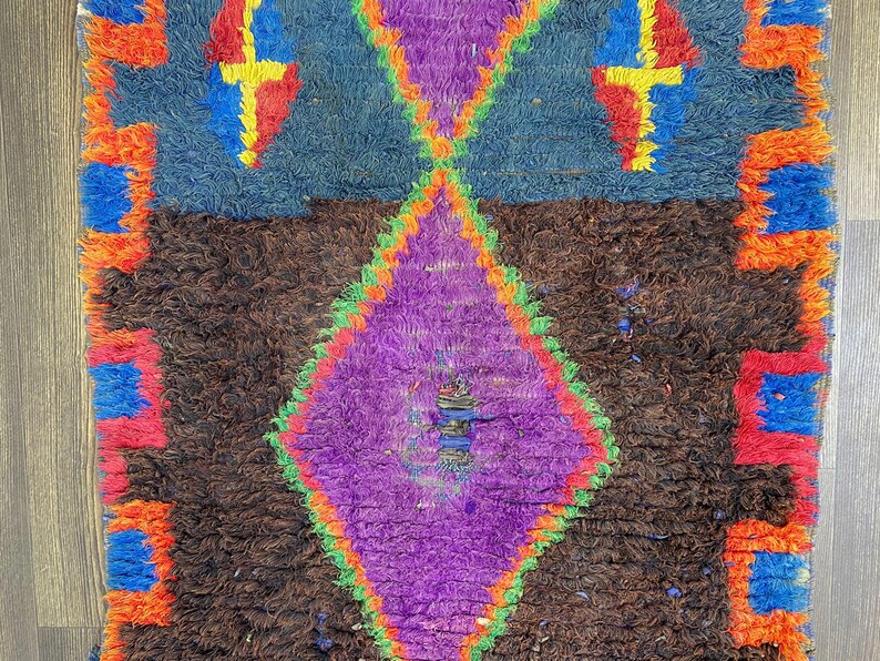 May include: A colorful, textured rug with a geometric design. The rug features a central purple diamond shape, framed by green and orange, set against a brown background. Blue and orange accents complete the design.