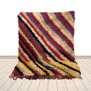 May include: A colorful, rectangular rug with a diagonal striped pattern. The rug features a variety of colors, including red, purple, black, yellow, and beige. The rug has a shaggy texture and a fringed edge.