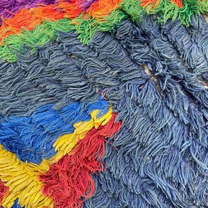 The image is a close-up of a textured fabric with a vibrant mix of colors, including shades of purple, green, orange, and yellow. The fabric appears to be made of yarn or thread, with a fuzzy, textured surface that creates a visually interesting and dynamic composition.
