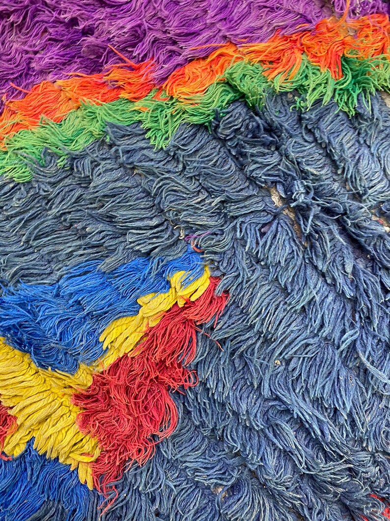 The image is a close-up of a textured fabric with a vibrant mix of colors, including shades of purple, green, orange, and yellow. The fabric appears to be made of yarn or thread, with a fuzzy, textured surface that creates a visually interesting and dynamic composition.