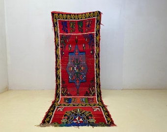 May include: A vibrant red rug with intricate geometric patterns in blue, green, and yellow. The rug features a central diamond design and decorative borders. The rug is displayed against a white wall and a light-colored floor.
