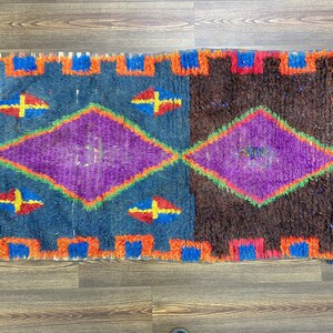 May include: A rectangular rug with a geometric design. The rug features a blue and brown background with purple diamond shapes and colorful accents. The edges are trimmed with orange and blue. The rug is placed on a wooden floor.