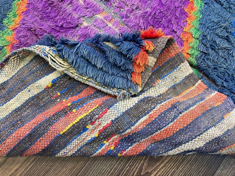 a close-up view of a colorful, textured rug with a mix of purple, blue, orange, and gray hues. The rug appears to be made of wool and has a distinctive, intricate pattern.