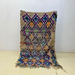 May include: A rectangular area rug featuring a beige base with a repeating diamond pattern in vibrant shades of purple, blue, orange, and green. The rug has a shaggy, textured pile and includes decorative fringe along the bottom edge.