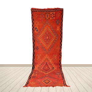 Peut inclure: Un long tapis rectangulaire de couleur rouge vif orné d'une série de grands motifs en losanges imbriqués dans des tons orange et brun foncé. La bordure est soulignée par un motif en zigzag noir, avec divers symboles géométriques dispersés.