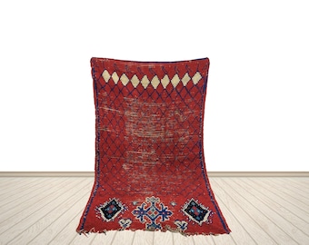 May include: A rectangular, red rug with a diamond pattern and a decorative border. The rug features a central diamond grid design in a darker red, with white and blue accents. The bottom border has geometric shapes in blue and white.