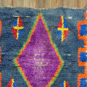 May include: A hand-woven rug with a central purple diamond design, outlined in green and orange, set against a blue background. The rug features geometric patterns in orange, blue, red, and yellow. The rug has a textured, shaggy appearance.