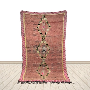 May include: A rectangular, hand-woven rug with a pink and black geometric design. The rug features a central pattern of diamond shapes in black, yellow, and pink. The border is a pink and yellow checkered pattern. The rug is displayed against a white background.