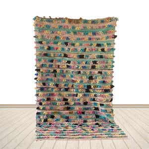 May include: A colorful, rectangular rug with horizontal stripes of various colors, including teal, pink, yellow, and black. The rug has a textured, woven appearance with a fringe along the top and bottom edges. The rug is displayed against a white background.