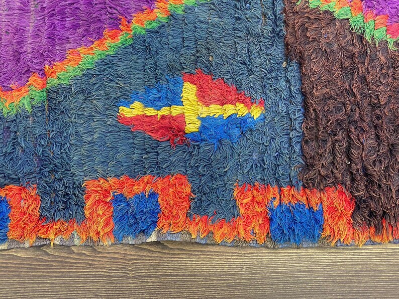 The image is a close-up of a colorful, textured rug with a geometric pattern. The rug has a mix of blue, red, and orange colors, and features a central design element that resembles a diamond shape. The rug appears to be made of wool or a similar material, and it is displayed on a wooden surface.