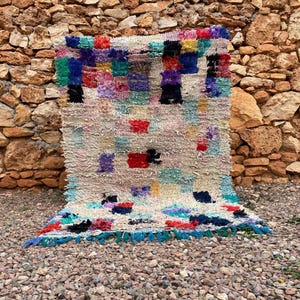 May include: A rectangular, hand-woven rug with a cream base and colourful, square patterns. The rug features red, blue, green, and purple. The edges are trimmed with a blue fringe. The rug is displayed against a stone wall, typical of Moroccan design.