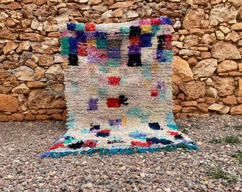May include: A rectangular, hand-woven rug with a cream-colored base and colorful, square-shaped patterns. The rug features a variety of colors, including red, blue, green, and purple. The edges are trimmed with a blue fringe. The rug is displayed against a stone wall.