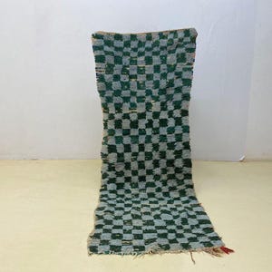 May include: A rectangular, hand-woven rug with a checkerboard pattern in shades of green and gray. The rug has a shaggy texture and a fringe at the bottom. The rug is displayed against a white wall.