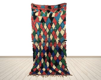 Può includere: Un tappeto colorato fatto a mano con un motivo a rombi. Il tappeto presenta un vivace mix di rombi rossi, blu, verdi, neri e color crema. Il tappeto è realizzato in un materiale tessuto e ha un bordo sfrangiato.