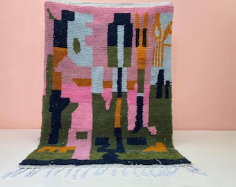 May include: A colorful, rectangular rug with an abstract design. The rug features a mix of pink, blue, green, orange, and navy blue geometric shapes. The rug has a fringe along the bottom edge and is suitable for home decor.