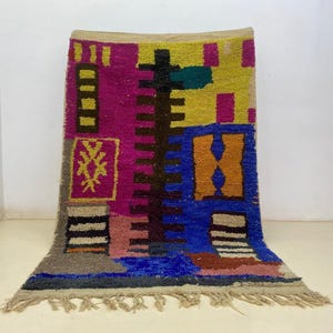 May include: A rectangular rug with a vibrant geometric pattern. The rug's design incorporates pink, yellow, blue, and brown hues. The rug has a fringe along the bottom edge, and measures approximately 150 cm in length.
