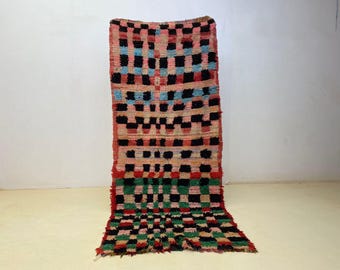 May include: A rectangular, hand-woven rug with a geometric pattern. The rug features a mix of colors, including pink, black, red, green, and blue. The design is a series of squares and rectangles, creating a textured look.
