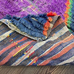 May include: A colorful, handmade rug with a mix of textures and patterns. The rug features a vibrant palette of purple, blue, orange, and green. The design includes striped sections and a textured, shaggy area. The edges are finished with blue fringe.