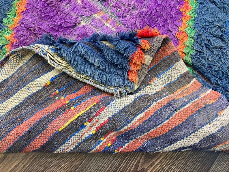 May include: A colorful, handmade rug with a mix of textures and patterns. The rug features a vibrant palette of purple, blue, orange, and green. The design includes striped sections and a textured, shaggy area. The edges are finished with blue fringe.