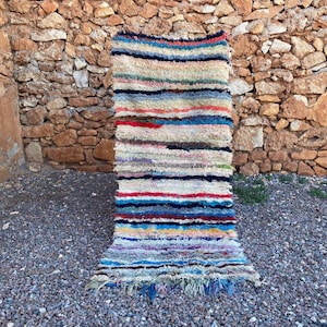 May include: A rectangular, striped rug with a textured appearance. The rug features horizontal bands of various colors, including blue, red, white, and yellow. The rug is displayed against a stone wall and gravel ground.