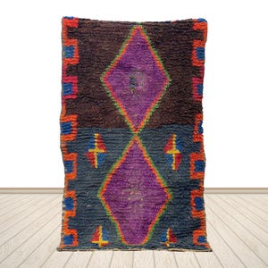 May include: A rectangular, hand-woven rug with a central diamond design in purple. The rug features a dark brown and blue background with orange, red, and blue geometric borders. The rug is displayed on a light wood floor.