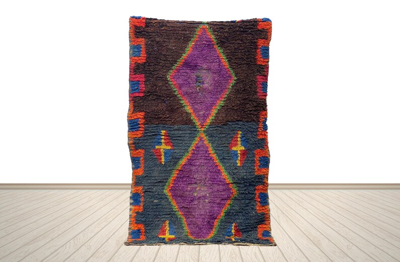May include: A rectangular, hand-woven rug with a central diamond design in purple. The rug features a dark brown and blue background with orange, red, and blue geometric borders. The rug is displayed on a light wood floor.
