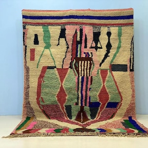 May include: A hand-woven rug with a beige background and colorful geometric patterns. The rug features diamond shapes in green and red, with vertical stripes in pink, blue, and brown. The top border has a striped design in various colors.