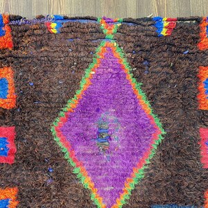 May include: A handmade rug with a brown background and a large purple diamond in the center. The diamond is outlined in green and red. The edges of the rug have blue, orange, and red accents.