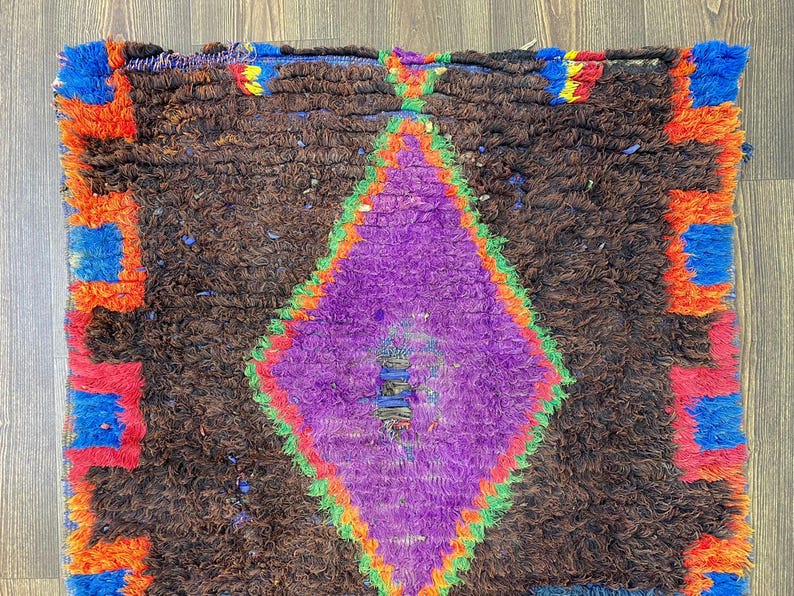 May include: A handmade rug with a brown background and a large purple diamond in the center. The diamond is outlined in green and red. The edges of the rug have blue, orange, and red accents.