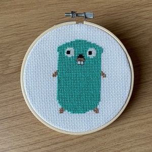 May include: A green embroidered gopher with brown paws and a white background, framed in a wooden hoop.
