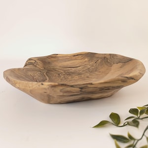 May include: A handcrafted wooden bowl with a natural, organic shape. The bowl is made from light brown wood with visible grain patterns and a smooth, polished finish. The bowl's irregular edges and shallow depth give it a rustic, artisanal feel.