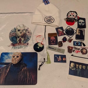 May include: A collection of Friday the 13th themed merchandise including a white drawstring bag with a Jason Voorhees graphic, a white beanie with a Camp Crystal Lake patch, a mousepad, a zippered pouch, various stickers, pins, and patches.