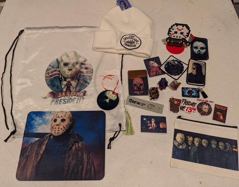 May include: A collection of Friday the 13th themed merchandise including a white drawstring bag with a Jason Voorhees graphic, a white beanie with a Camp Crystal Lake patch, a mousepad, a zippered pouch, various stickers, pins, and patches.