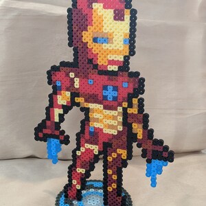 May include: A red and yellow Iron Man character made of perler beads, standing on a blue and black circular base.