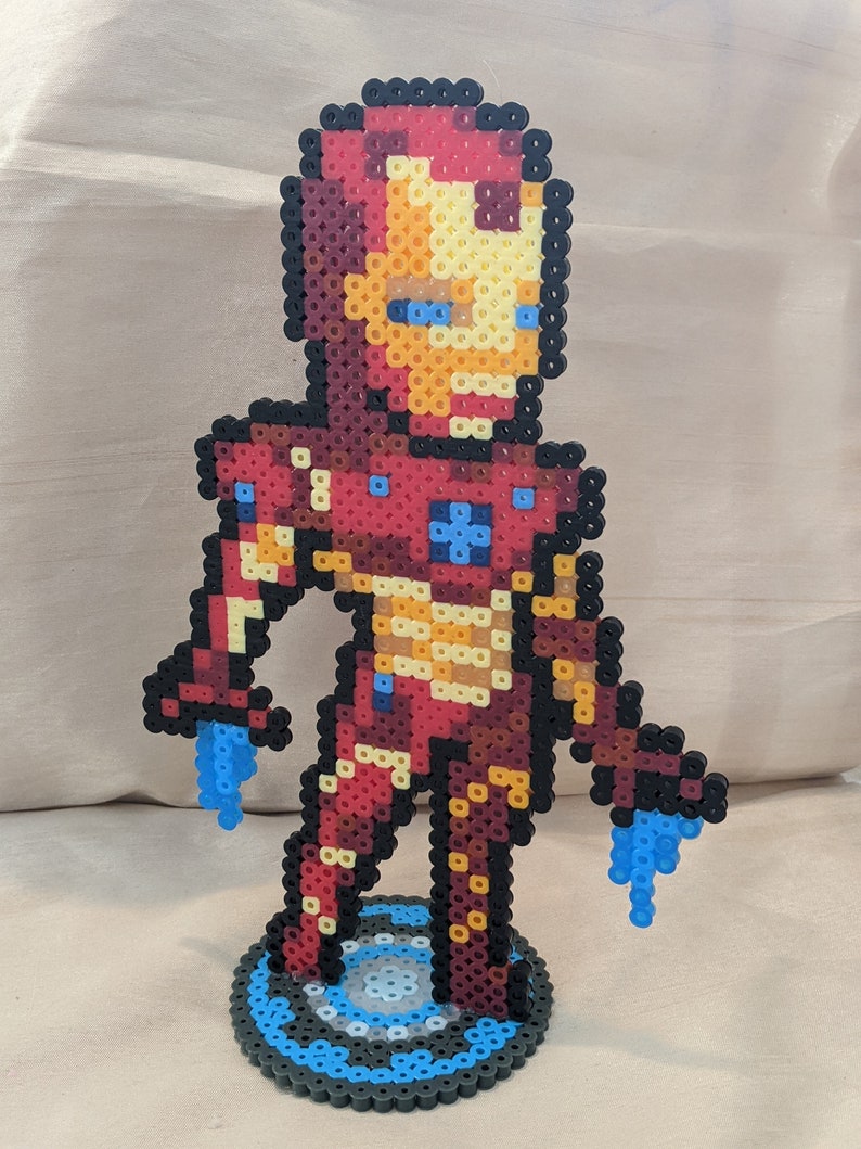 May include: A red and yellow Iron Man character made of perler beads, standing on a blue and black circular base.