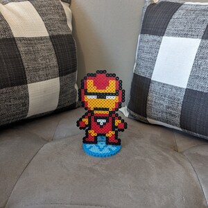 May include: A red, yellow, and white pixel art figure of Iron Man standing on a blue pixel art base. The figure is on a gray couch.
