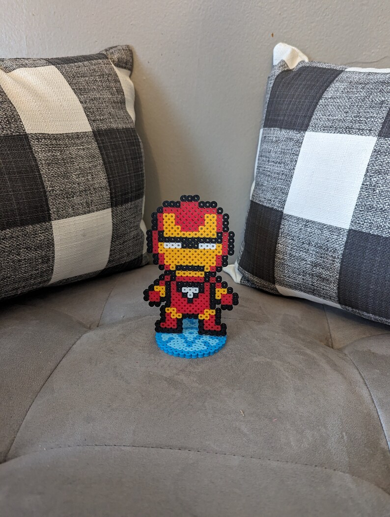 May include: A red, yellow, and white pixel art figure of Iron Man standing on a blue pixel art base. The figure is on a gray couch.