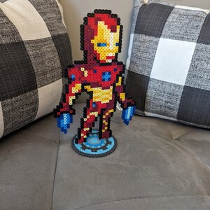 May include: A red, yellow, and blue pixel art figurine of Iron Man standing on a blue and silver pixel art base.
