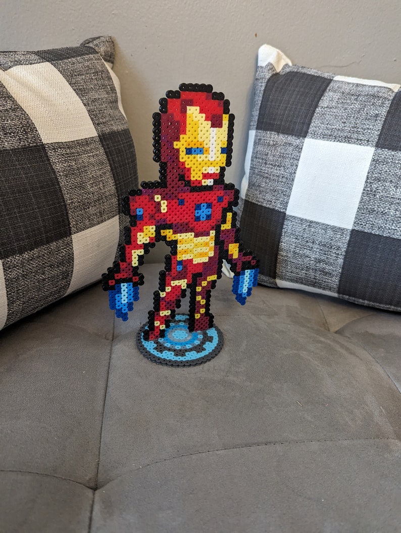 May include: A red, yellow, and blue pixel art figurine of Iron Man standing on a blue and silver pixel art base.
