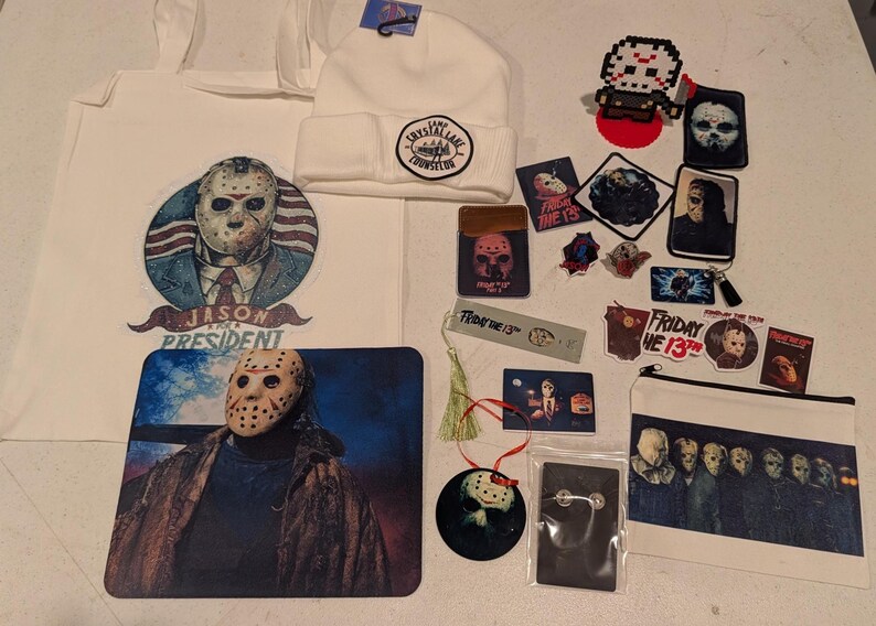 May include: A collection of Friday the 13th merchandise including a white tote bag with a Jason for President graphic, a white beanie with a Camp Crystal Lake patch, a mousepad, stickers, a bookmark, a zipper pouch, and various pins and patches.