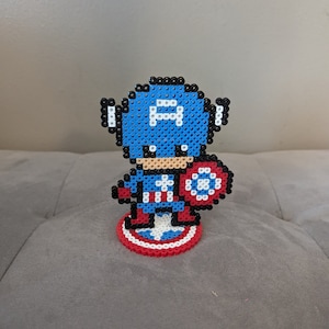 May include: A blue and red pixel art figurine of Captain America holding his shield. The figurine is standing on a red, white, and blue pixel art base.