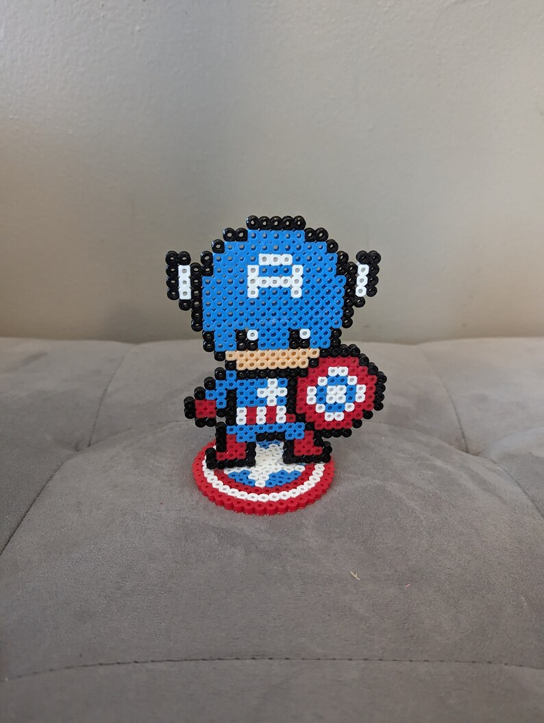 May include: A blue and red pixel art figurine of Captain America holding his shield. The figurine is standing on a red, white, and blue pixel art base.
