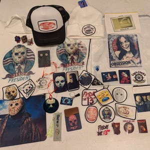 May include: A collection of Friday the 13th themed merchandise including a black and white trucker hat, a white beanie, a tote bag, a drawstring bag, various stickers, patches, pins, a bookmark, and small pouches featuring Jason Voorhees imagery.
