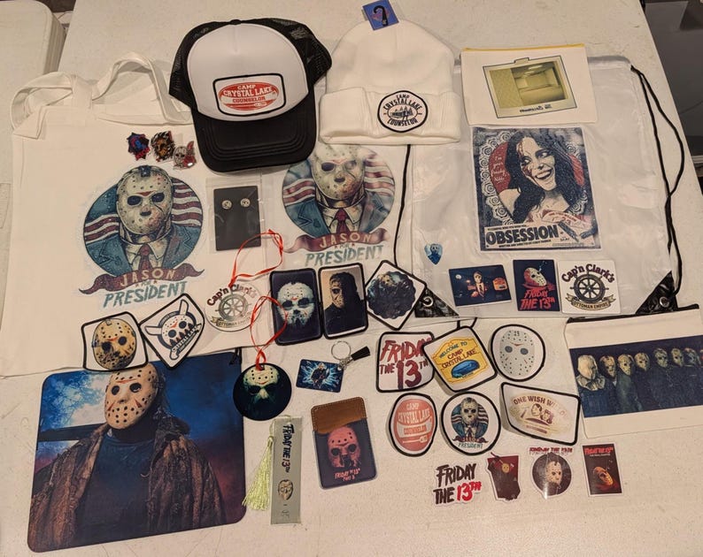 May include: A collection of Friday the 13th themed merchandise including a black and white trucker hat, a white beanie, a tote bag, a drawstring bag, various stickers, patches, pins, a bookmark, and small pouches featuring Jason Voorhees imagery.